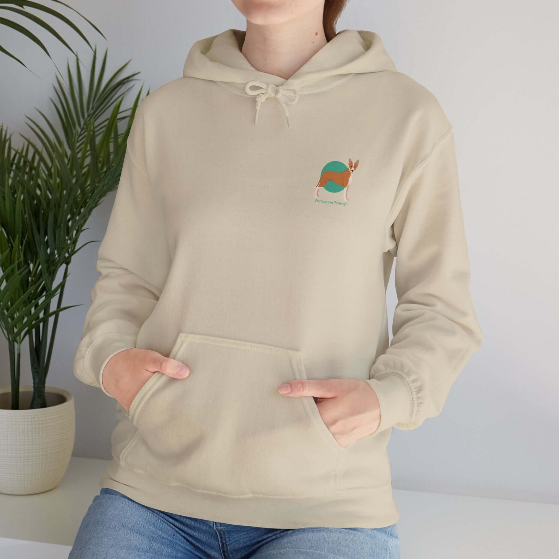 Woman wearing a beige hooded sweatshirt featuring a small embroidered Portuguese Podengo dog design, standing indoors near a potted plant.