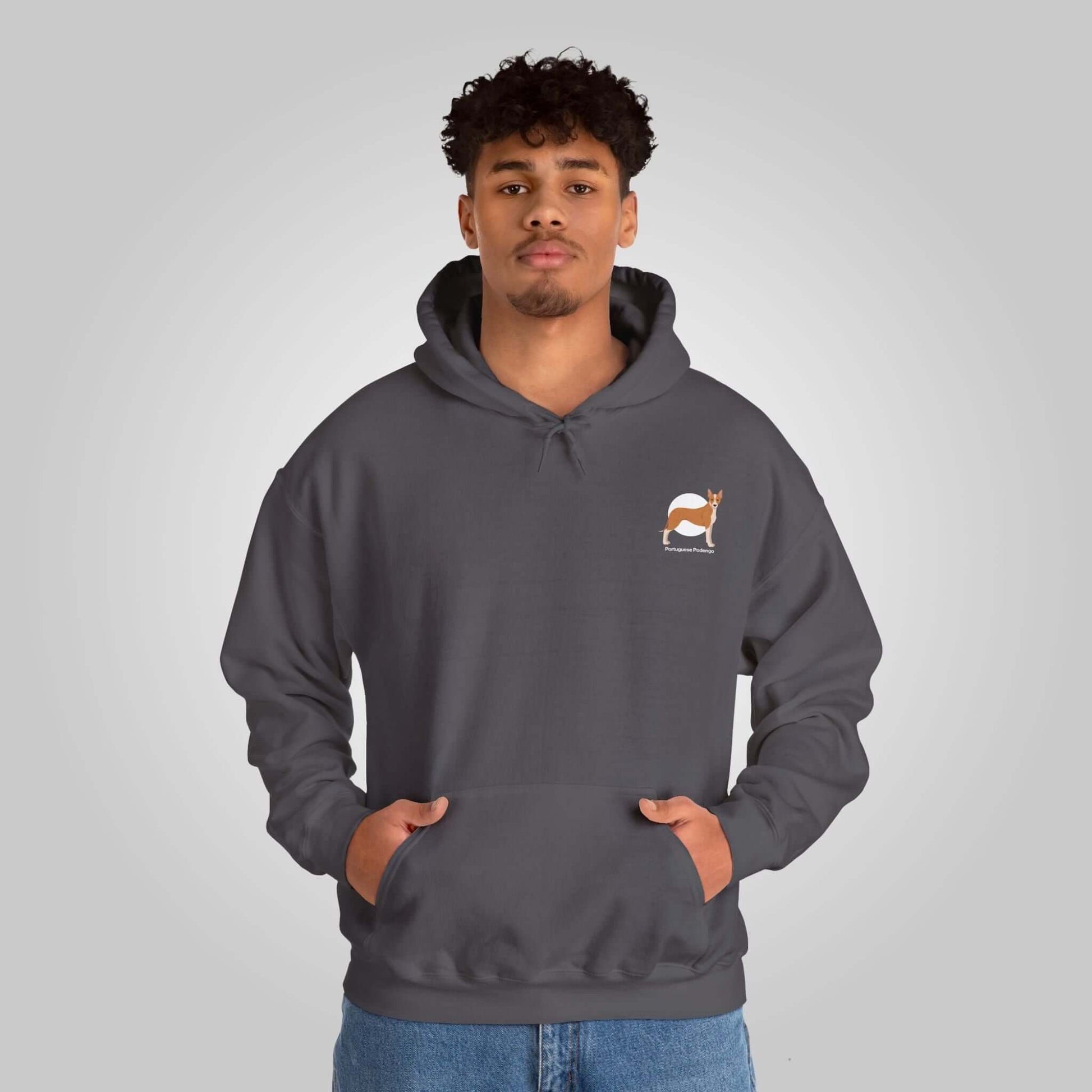 Young man modeling a Portuguese Podengo unisex hoodie in dark gray with dog logo, hands in pockets, standing against a neutral background.