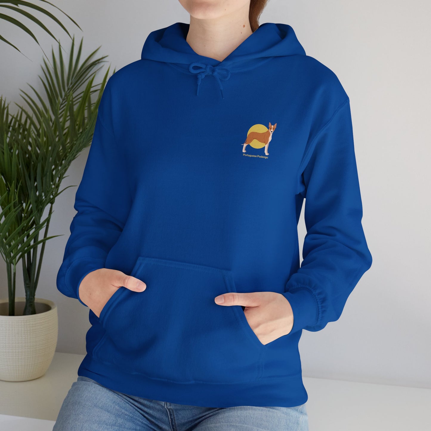 Woman wearing a blue hoodie featuring a small Portuguese Podengo dog design, standing indoors near a potted plant.