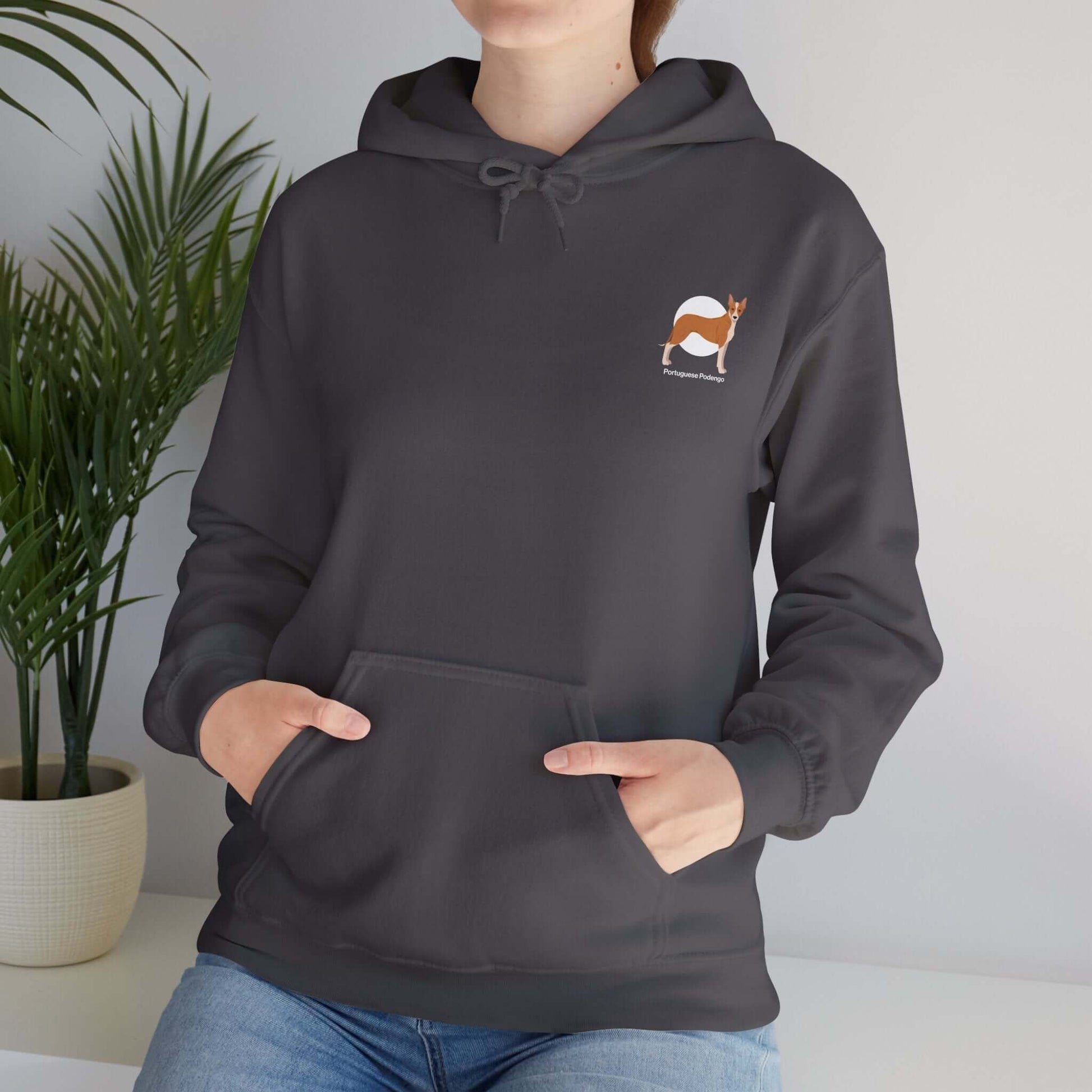 Woman wearing Portuguese Podengo dog hoodie in dark gray, hands in pocket, with plant in background.